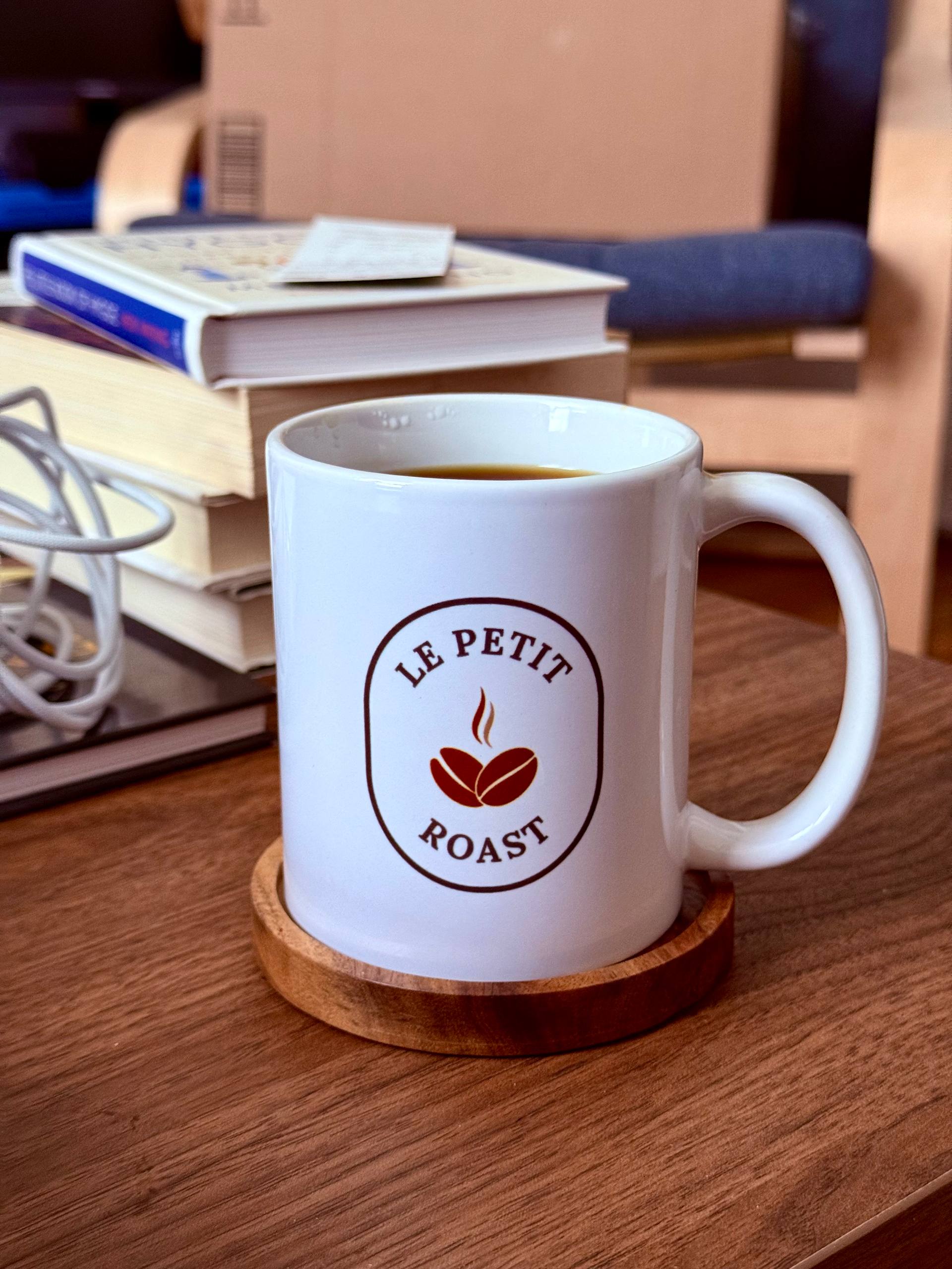 White ceramic mug with Le Petit Roast logo filled with fresh coffee, sitting on a wooden coaster on a desk with books in the background